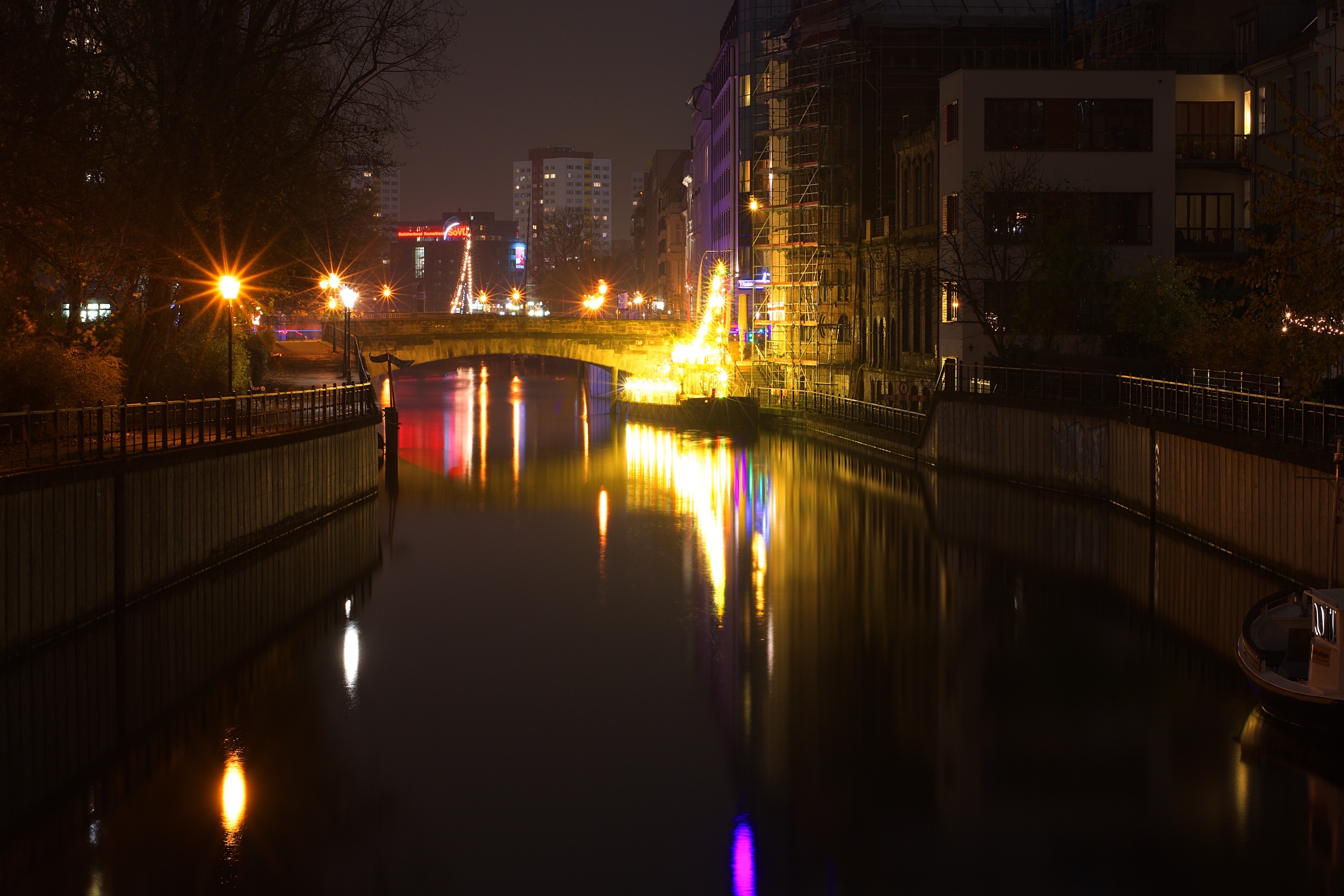 picture of a river in the centre of the frame at night. There is  construction scaffolding on one building to the right, a tall apartment or  office building in the distance, and the surrounding lights are all  reflected in the river-water.