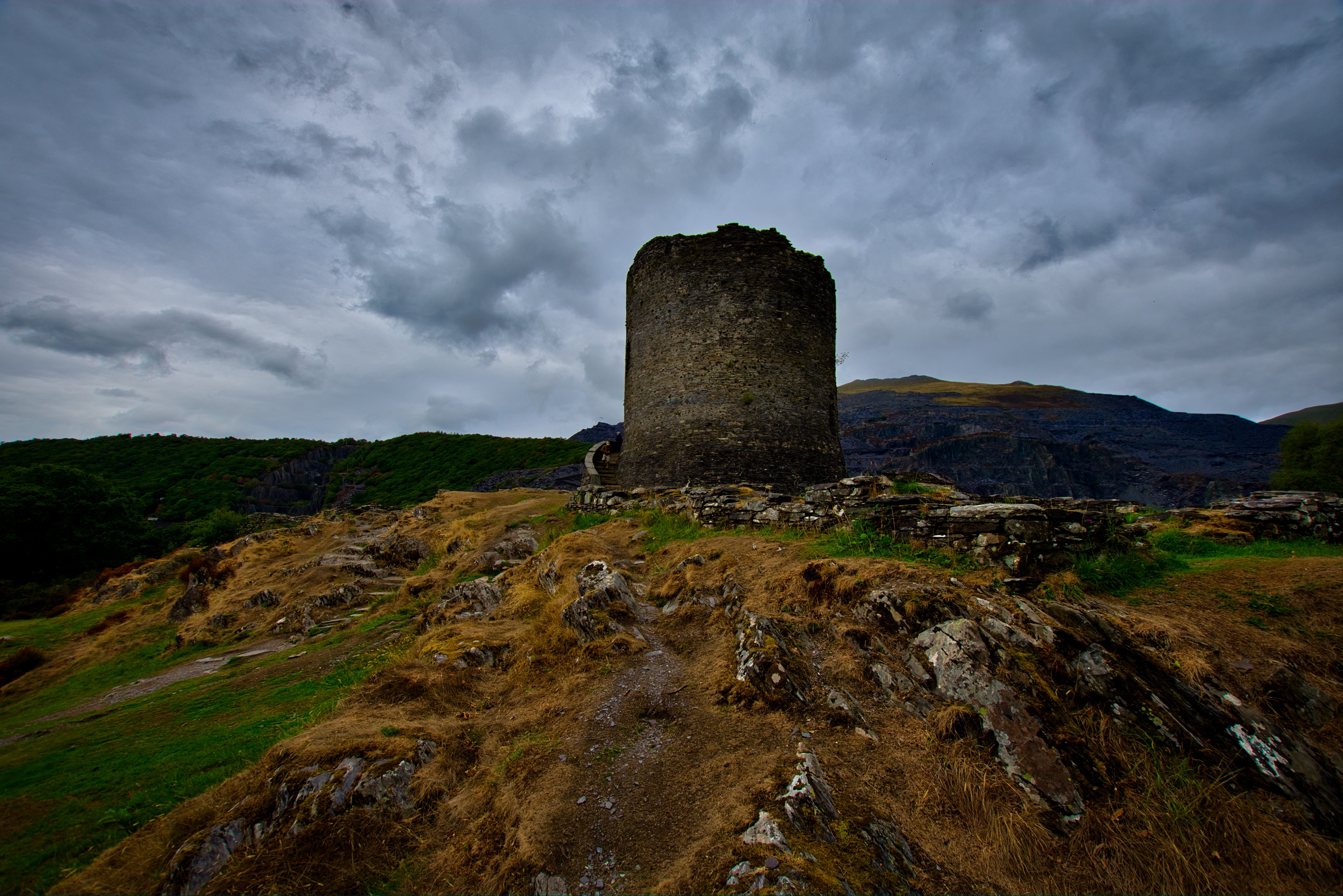 A ruined tower in the centre of the shot, clearly missing the top portion.  In front of the tower is orange-ish tussock through which rocks protrude,  and the remains of the walls that once surrounded the tower. The sky is  overcast, and the whole area looks dark, as though it has just or is about  to rain.